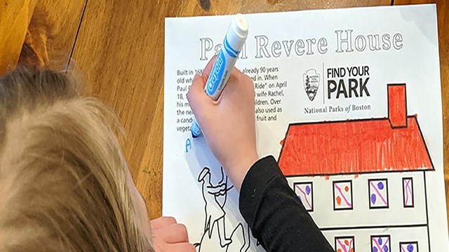 A young girl with blonde hair colors a picture of the Paul Revere House on a wooden table.