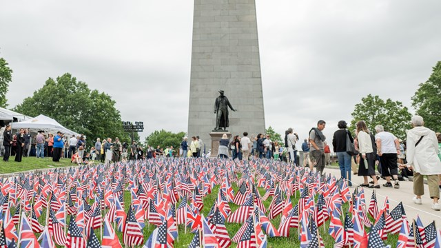 People surround a flag garden of American and British flags in front of the Bunker Hill Monument