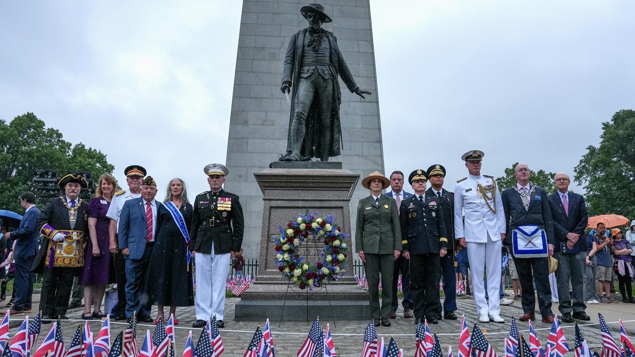 dignitaries in suits or uniforms stand around the Warren Prescott statue with a wreath.