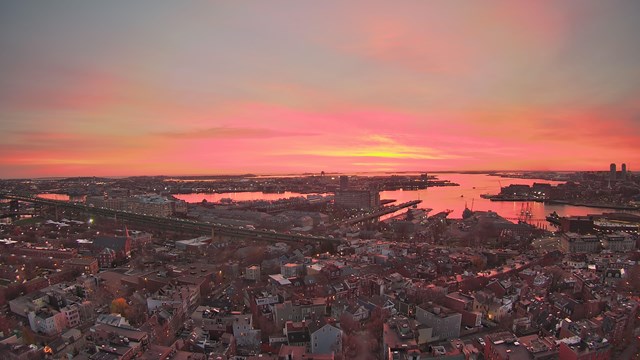 Web camera view of downtown Boston and Boston Harbor at sunset. 