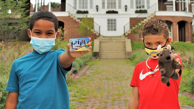 Two kids wearing masks standing side-by-side, one holds up a picture, another has a stuffed bison.