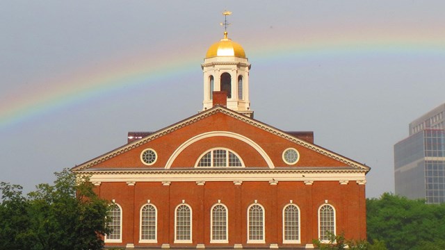 Faneuil Hall stands stoically after a rainstorm, with a rainbow in the background.