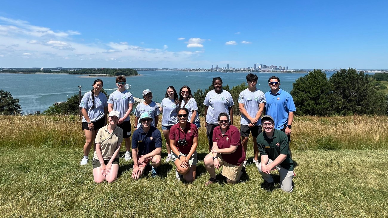 A group of youth wearing gray tshirt uniforms with water and the Boston skyline in the background