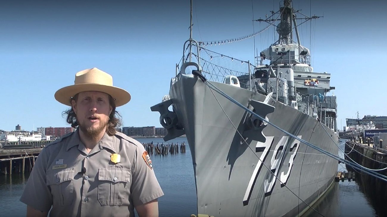 An NPS ranger in uniform standing in front of the bow of a destroyer in water. 