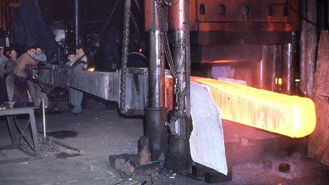 An anchor being forged in a shop with the end of metal bright yellow & men working at the other end.