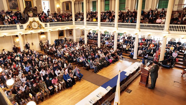 person on stage speaking from a podium to a crowded two-story meeting hall.