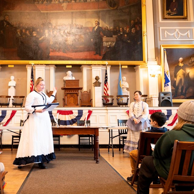 three adults stand in front of the stage at Faneuil Hall as students look on.