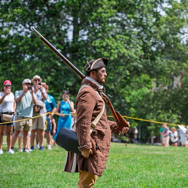 A Park ranger dressed in period clothing has a musket on his shoulder in front of an audience