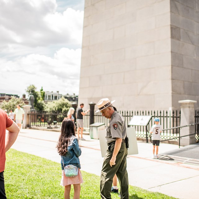 A young kid listening to a Ranger speak in front of a granite monument.