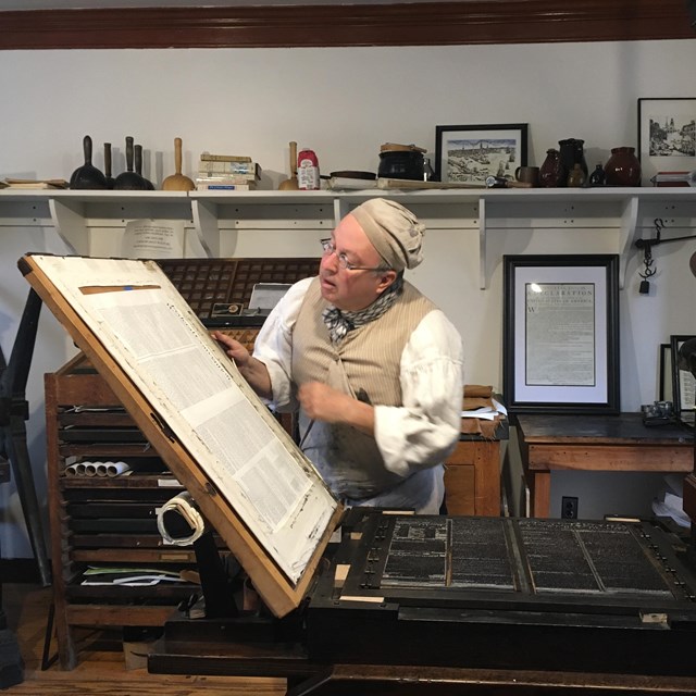 printer in historical clothing examining a freshly printed page on the press.