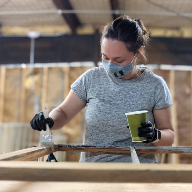 A historic preservation corps employee paints a wooden window frame, holding a brush and paint can.