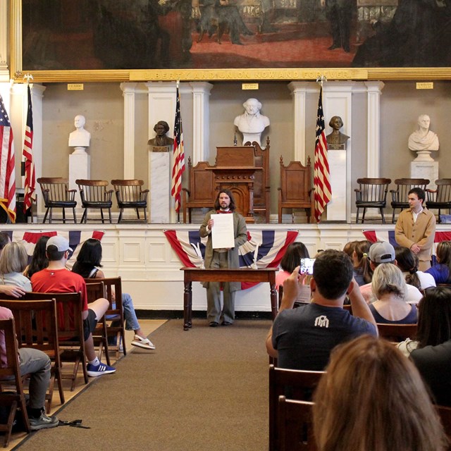 Interior of a large hall with rangers in 1800s clothing speaking to a sitting audience. 