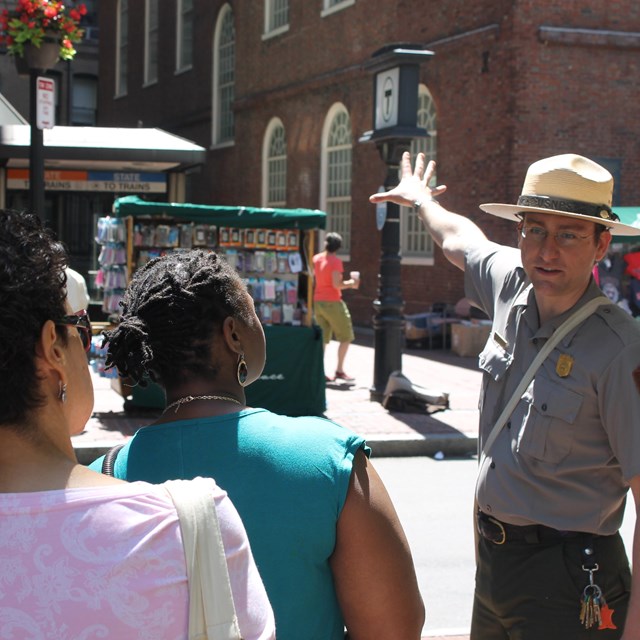 A park ranger hand raised towards a historic building speaking to a diverse tour group.