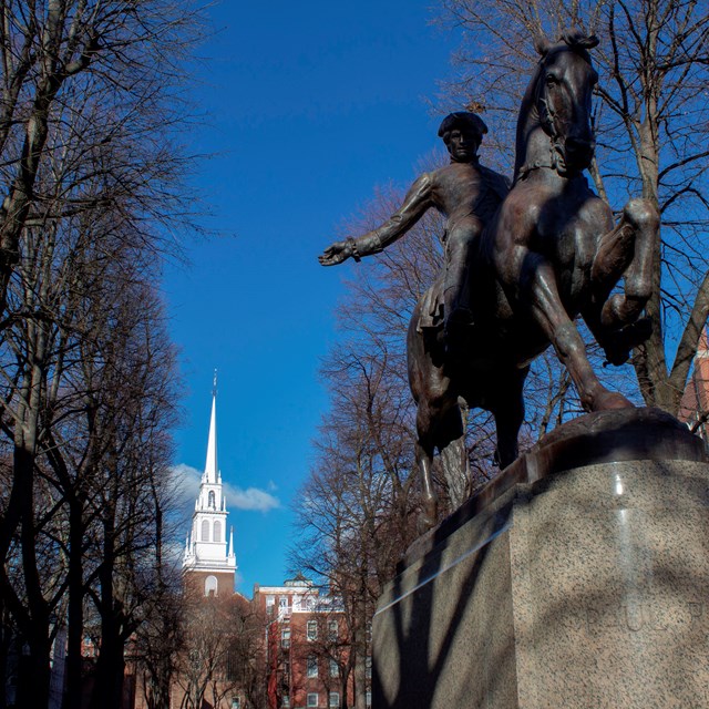 Statue of a man on horseback waving his arm. In the background is a white steeple.