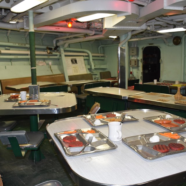 The mess of the USS CASSIN YOUNG which has white square tables with gray food trays on top. 