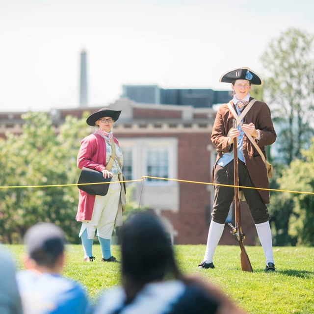 Two Park rangers dressed in period clothing, one has a musket to her side and speaks to the audience