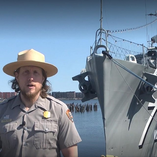 An NPS ranger in uniform standing in front of the bow of a destroyer in water. 