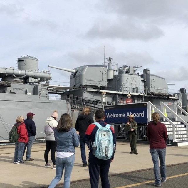 spread out groups of people facing a ranger giving a talk. Naval destroyer in the background.