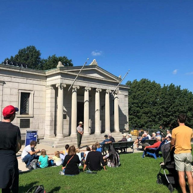 People standing and sitting on grass listening to a Ranger speak in front of a granite building.