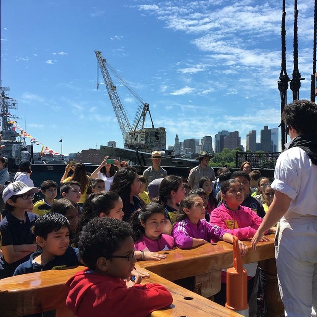 Students aboard the USS Constitution gathered around a sailor who is speaking to them.