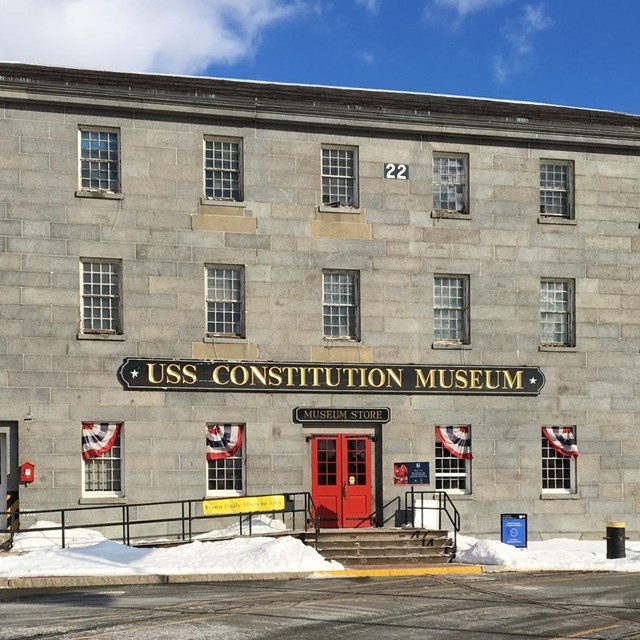 A grey stone 3-stroy rectangular building with a red door in the center and museum sign above.
