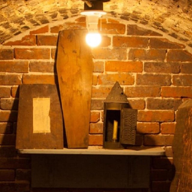 Inside a small red-brick crypt with a shelf that has a couple boxes and a coffin shaped box.