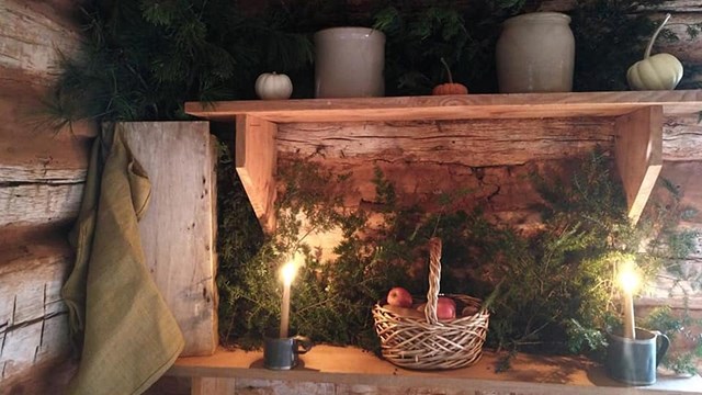An interior scene of a cabin and a wooden table with pine boughs, candles and an apple basket. 