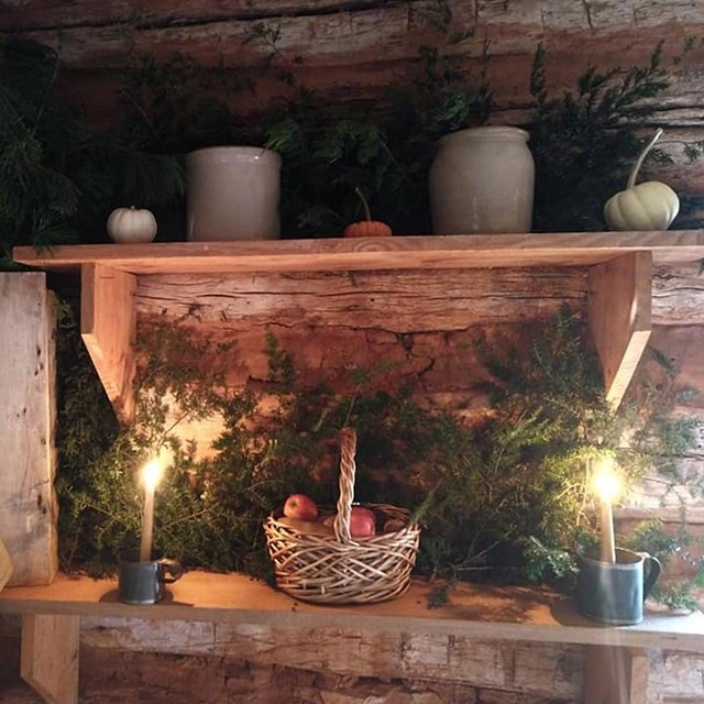 An interior scene of a cabin and a wooden table with pine boughs, candles and an apple basket. 