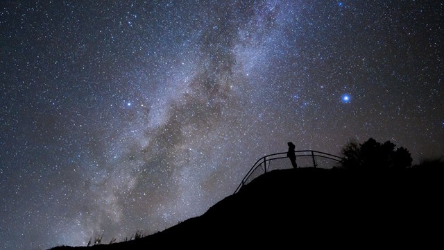 A dark sky with lots of stars with a person standing behind a railing in the foreground.