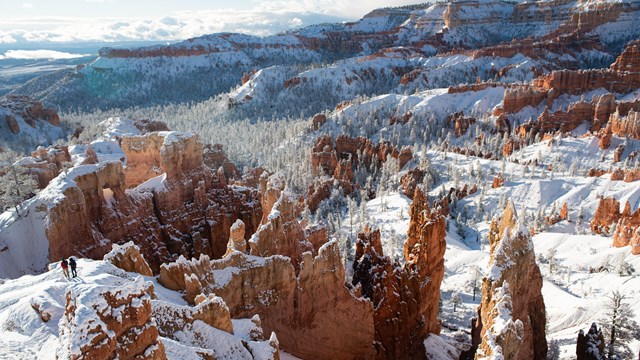 Two hikers walk amongst snow covered red rock formations in an amphitheater 