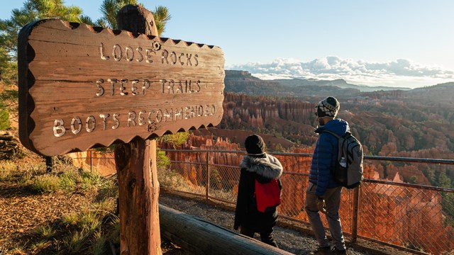 Two hikers walk past a sign that says Loose Rocks Steep Trails Boots Recommended