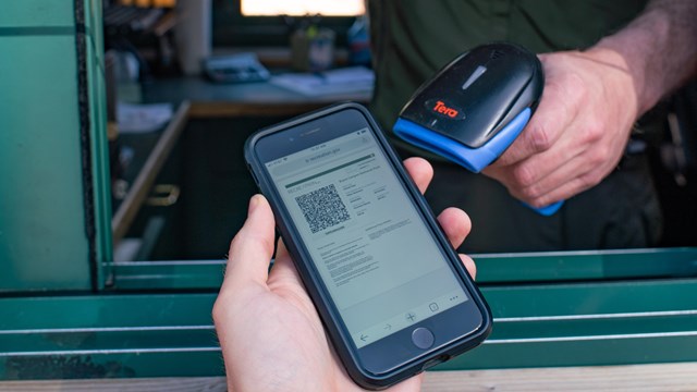 A man holds up a cell phone as another man in a booth holds a scanner to the phone.