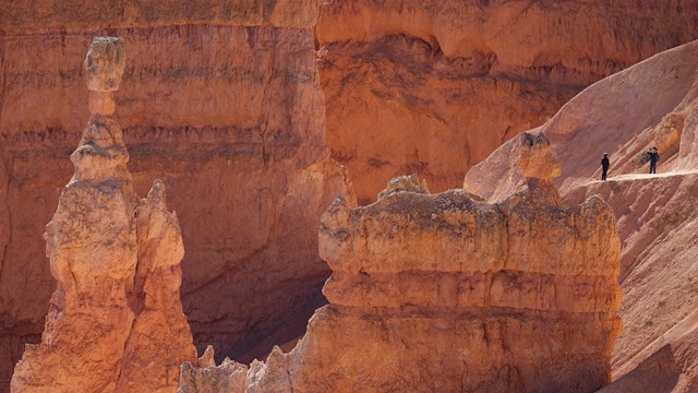 Two hikers walk amongst red rock formations with Thor's Hammer visible on the left.