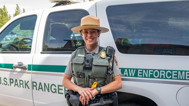 A law enforcement park ranger stands in full uniform in front of her white and green patrol vehicle.