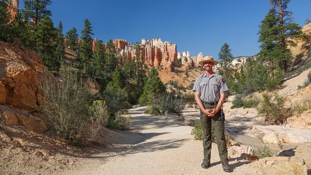 A park ranger in uniform stands on a path in front of red rock formations in the background.