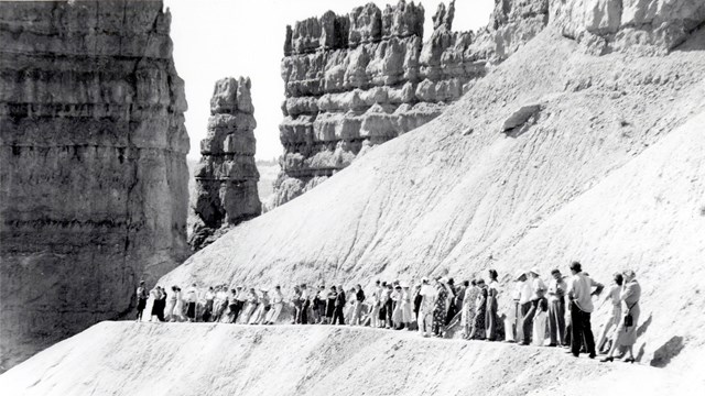 A black and white photo of a group of people along a narrow trail below tall rock spires