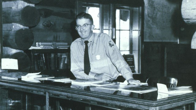 A black and white photo of a park ranger in uniform standing behind a counter.