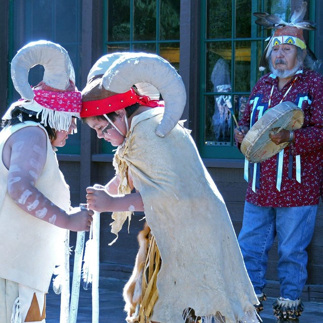 Two children with mountain sheep garb butt heads while elder plays drum in background
