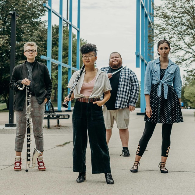 Six people stand in paved plaza holding instruments