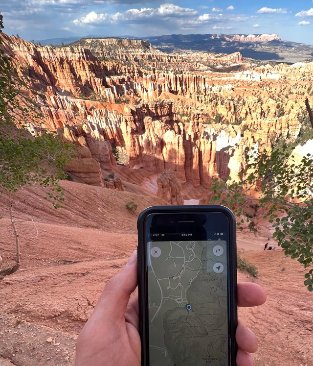 Hand holds a smartphone showing a map in front of a vast landscape of red rock spires and cliffs.