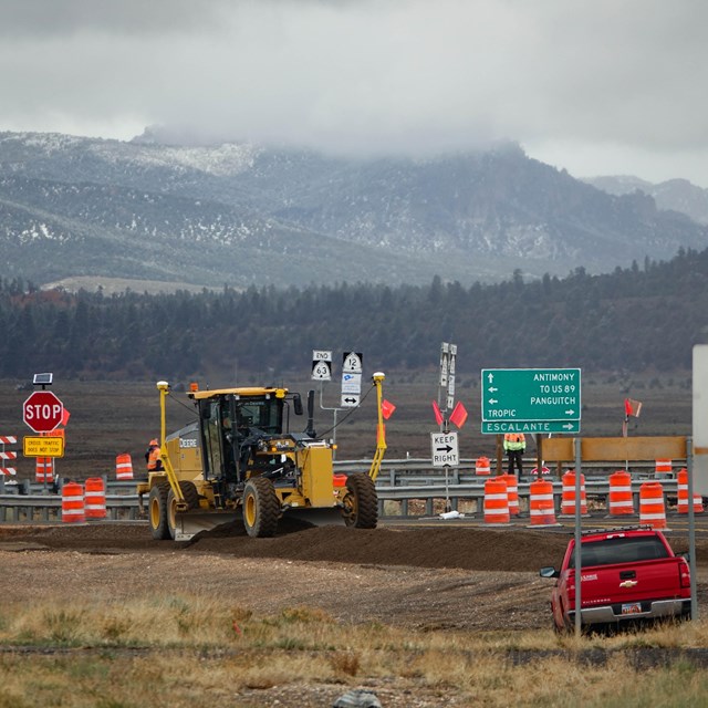 construction zone with tractors, vehicles, road signs, and cloudy mountains and forest beyond