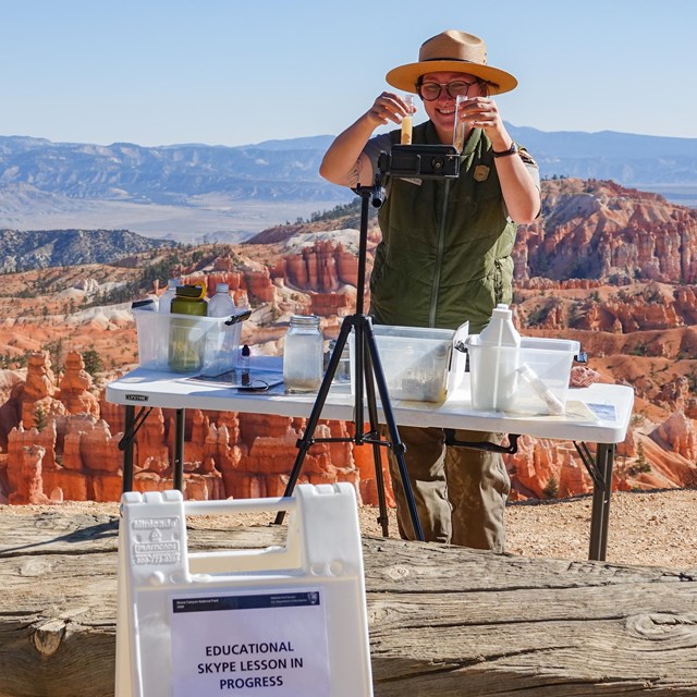 Woman in park ranger uniform stands showing test tubes as part of virtual program