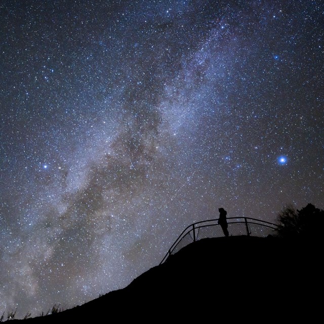 A dark sky with lots of stars with a person standing behind a railing in the foreground.