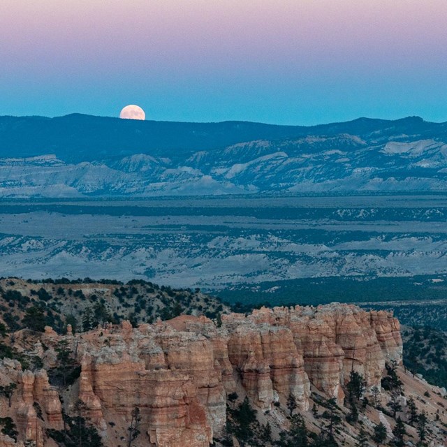 A full moon rises behind a mountain in a pink and blue sky.