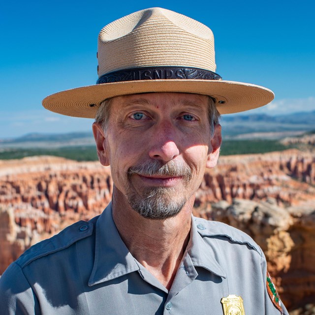 Man stands with straw park ranger hat, grey uniform shirt, nametag, and golden badge before vast red