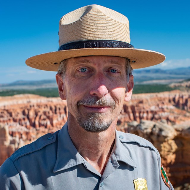 Man stands with straw park ranger hat, grey uniform shirt, nametag, and golden badge before vast red