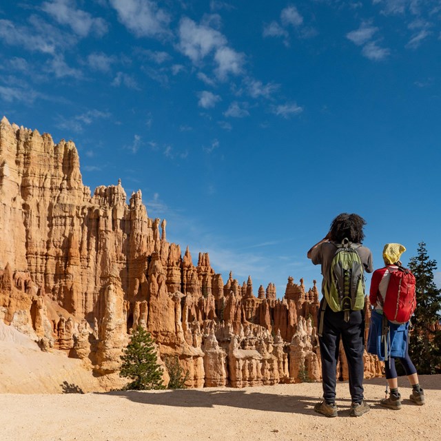 A couple stands along a trail beneath red rock spires and tall limestone cliffs.