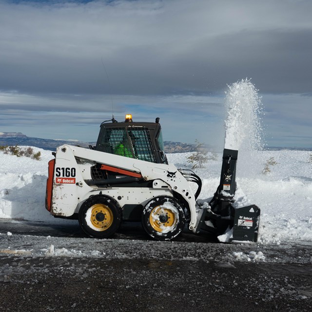 A snowplow blows snow along a roadside.