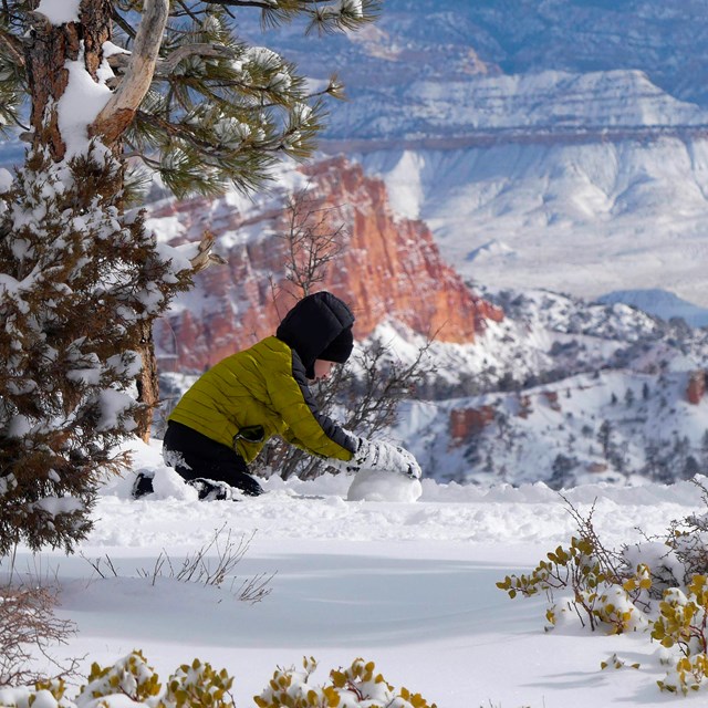 A boy in winter clothing rolls snow into a small ball along a cliff edge.