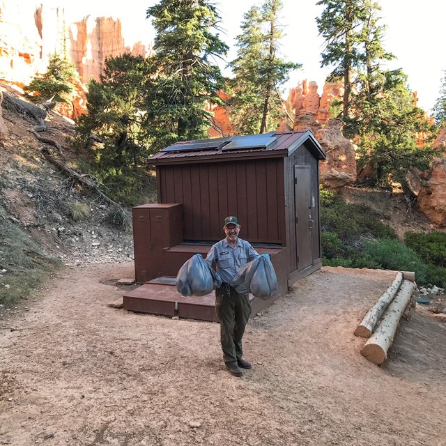 Man in park maintenance uniform stands holding two grey bags in front of brown outdoor toilet